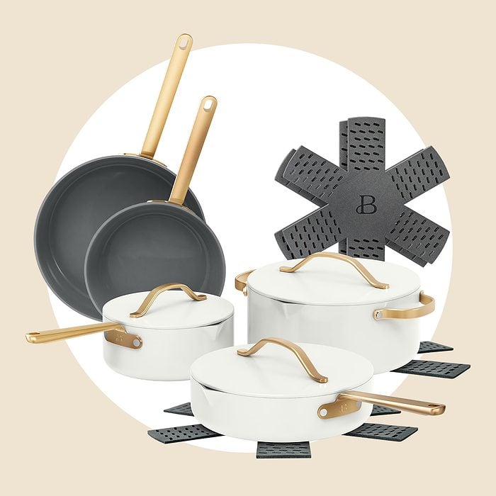 A set of white cookware with gold handles, including two frying pans, a saucepan, a sauté pan, and a Dutch oven, arranged with gray pan protectors underneath each piece, on a beige background.
