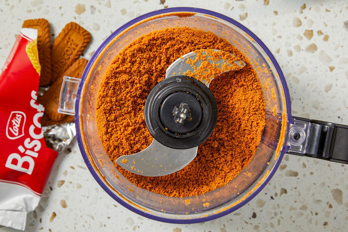 Overhead horizontal image of crushed Lotus Biscoff cookies in a food processor. An open package of Lotus Biscoff cookies is to the left of the processor.