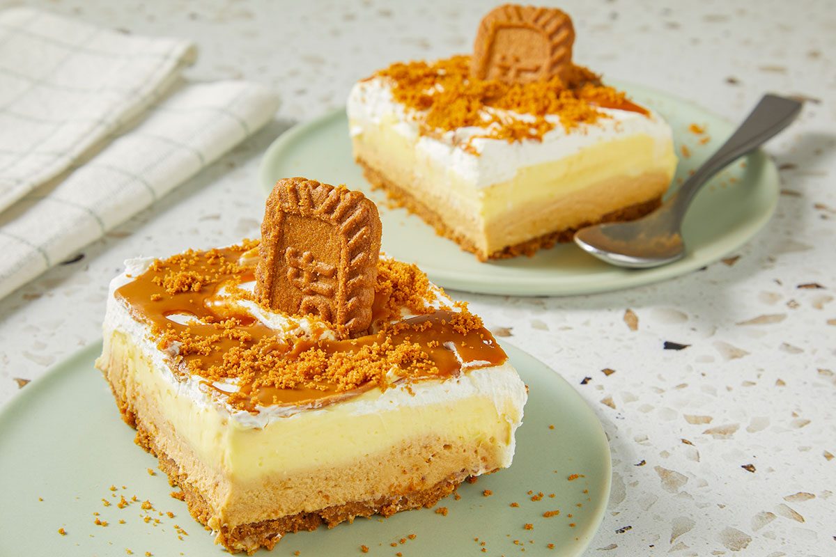 Eye Level horizontal image of two slices of Lotus Biscoff cookie cheesecake. The cake has a cookie crust, layers of different creams, and is decorated with caramel drizzle, cookie crumbs, and a Lotus Biscoff cookie.