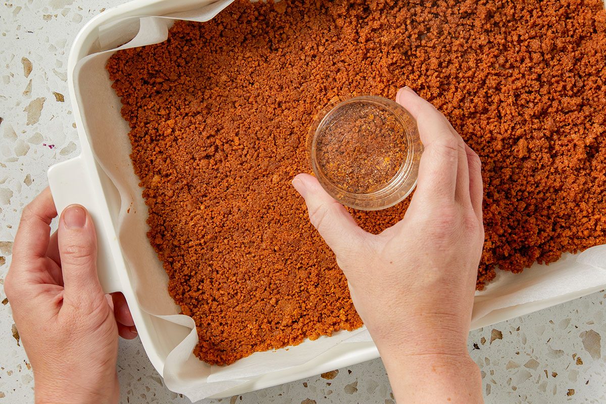 Overhead horizontal image of a graham cracker crust being pressed into a white baking dish lined with parchment paper. A small glass is being used to pack down the crust.