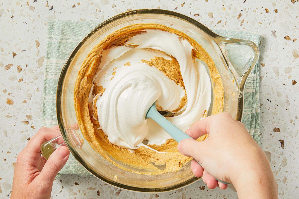 Overhead horizontal image of a person using a blue spatula to fold whipped egg whites into a tan batter within a clear glass mixing bowl. A green and white checkered towel sits to the left of the bowl on a countertop.
