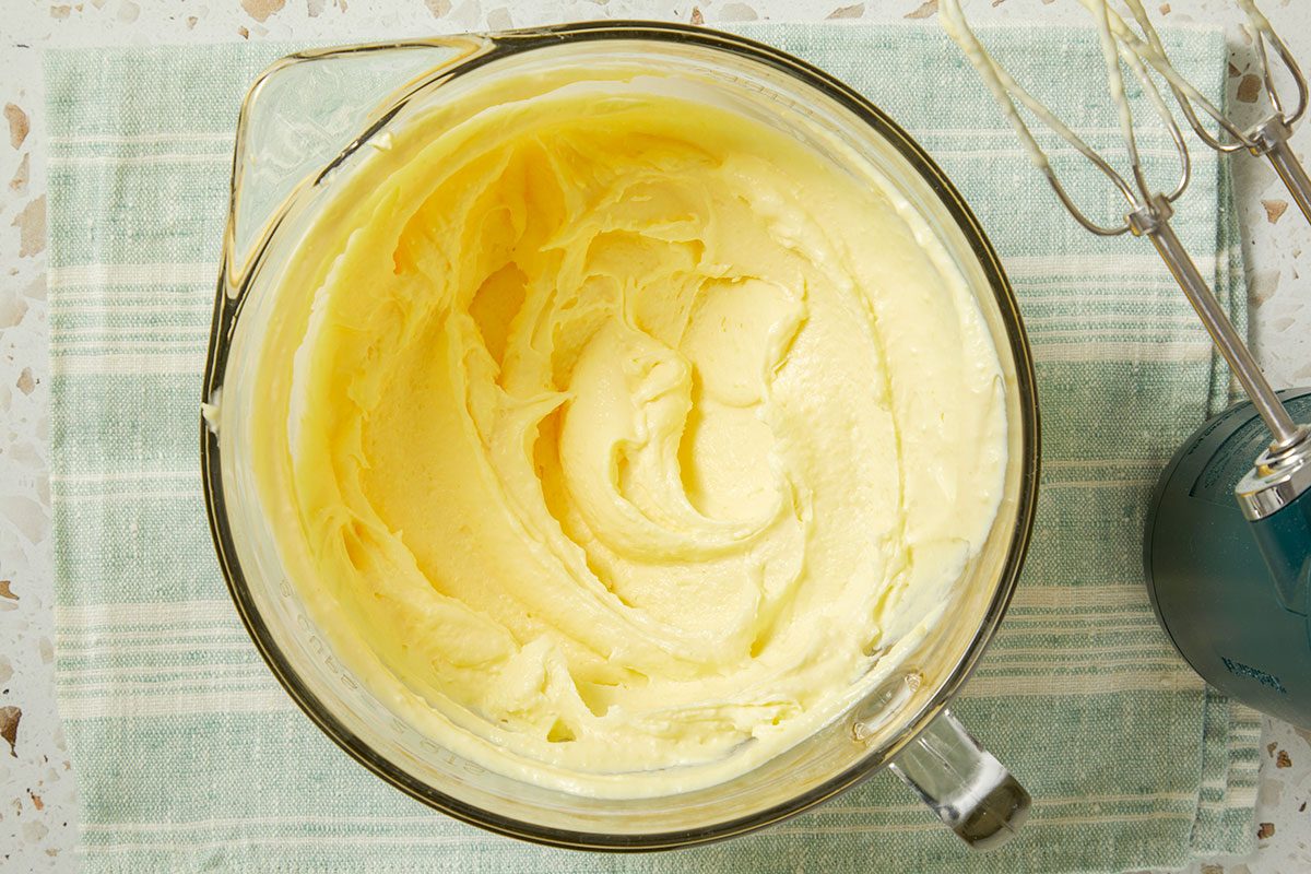 Overhead horizontal image of the batter in a glass measuring bowl after using an electric mixer. The light yellow batter has been mixed well and sits inside the clear bowl next to the electric mixer with beaters attached.