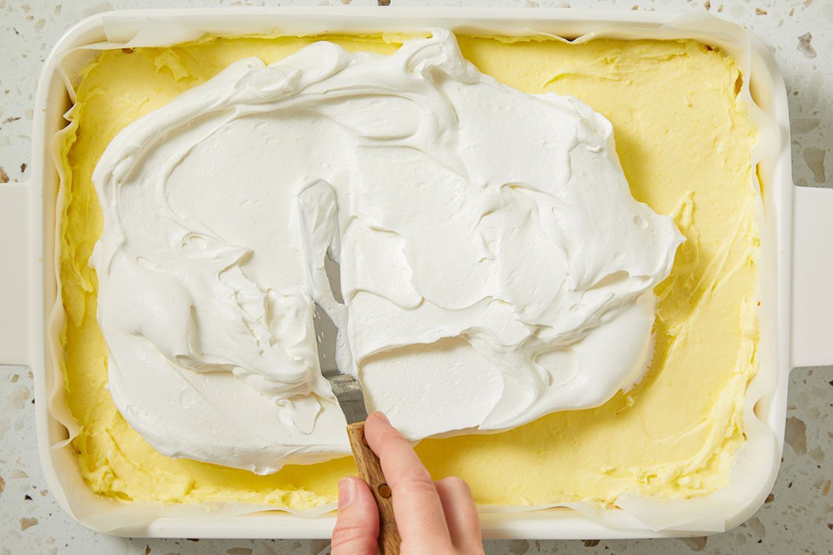 Overhead horizontal image of a baker using a metal spatula to spread white frosting over a yellow cake in a white baking dish lined with parchment paper. The dessert is placed on a speckled countertop.