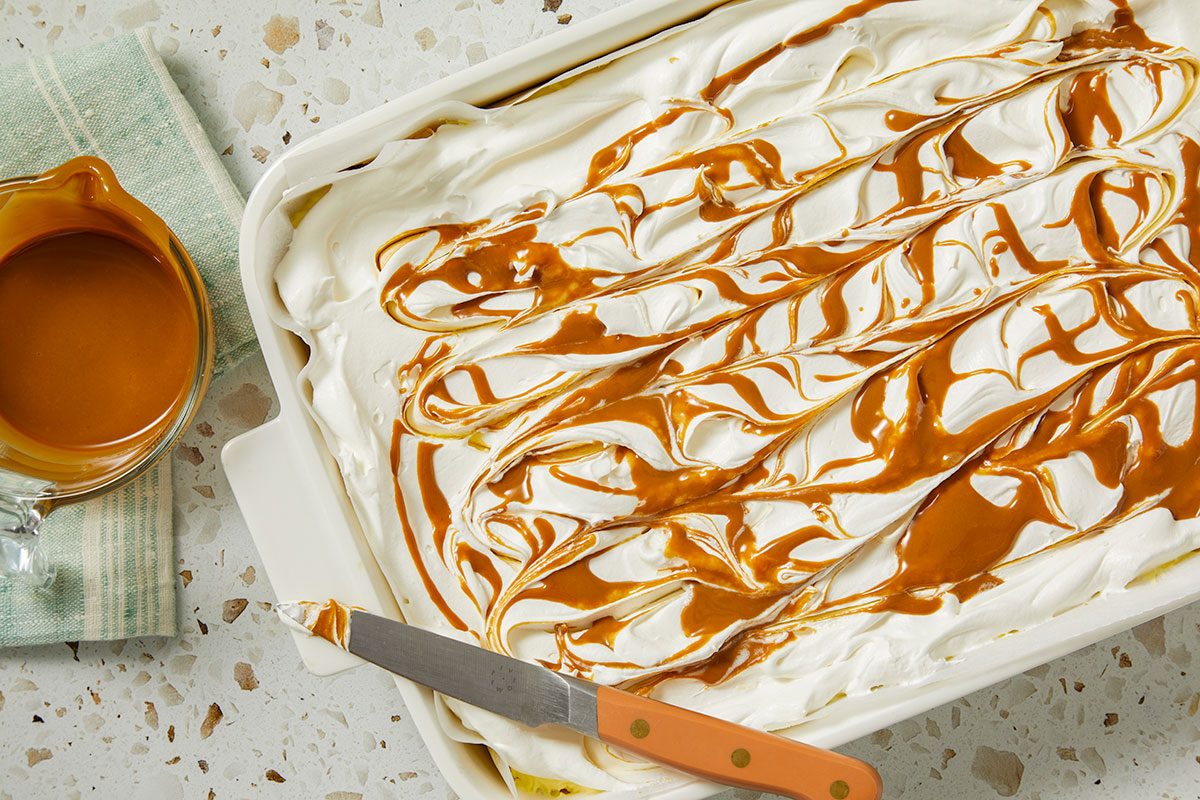 Overhead horizontal image of a dessert in a white baking dish being drizzled with caramel. The top layer of the dessert is a creamy white color, and the caramel is drizzled across the top in wavy lines. A glass measuring cup filled with caramel and a knife are also present.