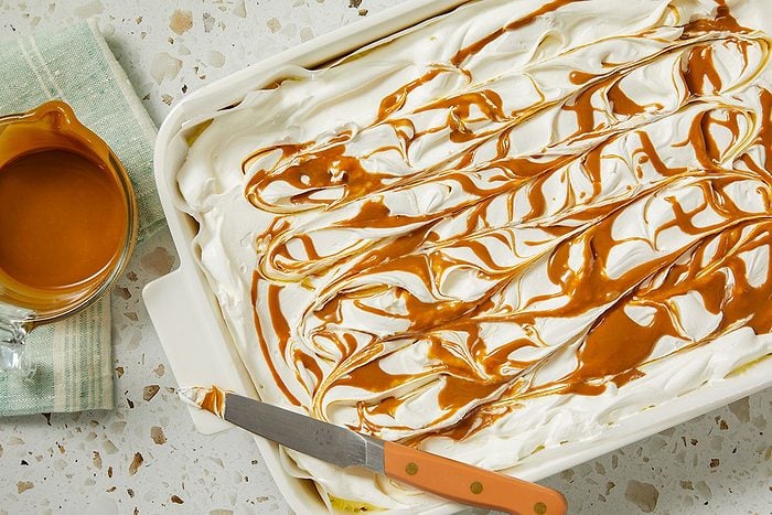 Overhead horizontal image of a dessert in a white baking dish being drizzled with caramel. The top layer of the dessert is a creamy white color, and the caramel is drizzled across the top in wavy lines. A glass measuring cup filled with caramel and a knife are also present.
