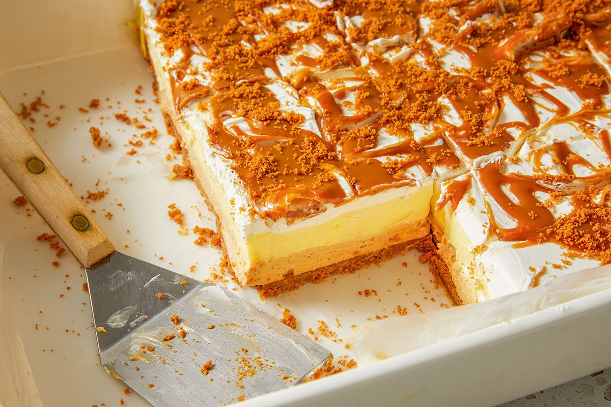 Overhead horizontal image of dessert bars in a white baking dish, one portion of the dessert removed with a metal spatula. The dessert bars feature a layered appearance, topped with caramel drizzle, whipped cream, and crushed cookies.
