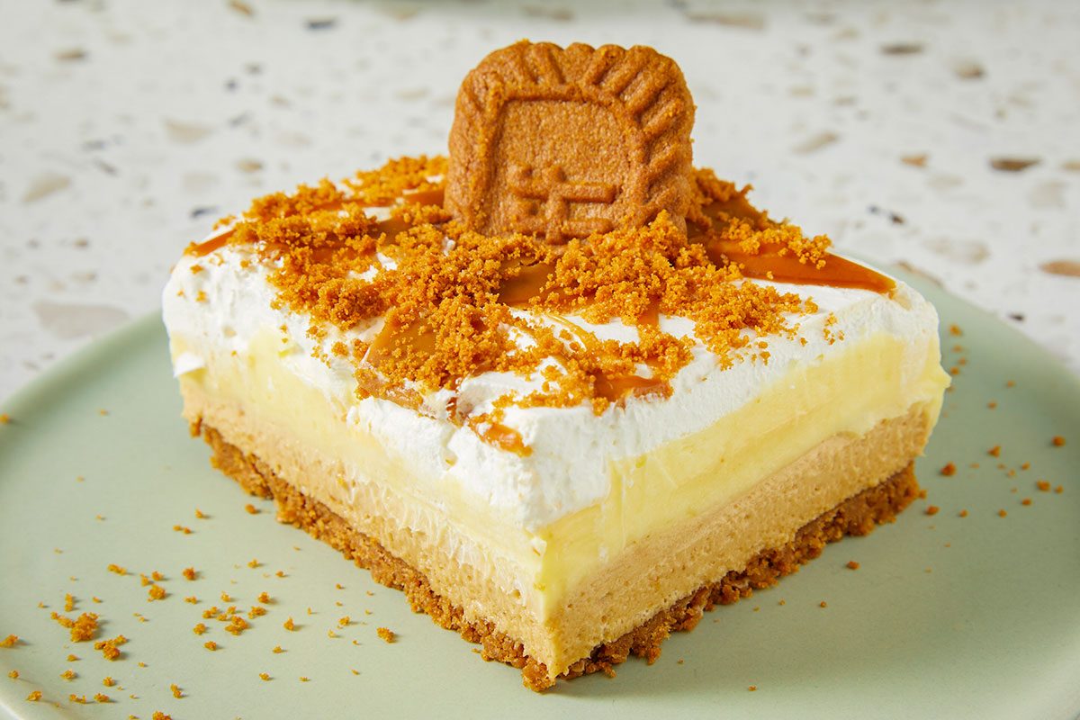 Eye Level horizontal image of a slice of layered dessert square on a green plate. The dessert has a crust, a filling, whipped cream and is topped with cookie crumbs, caramel, and a whole Lotus Biscoff cookie.