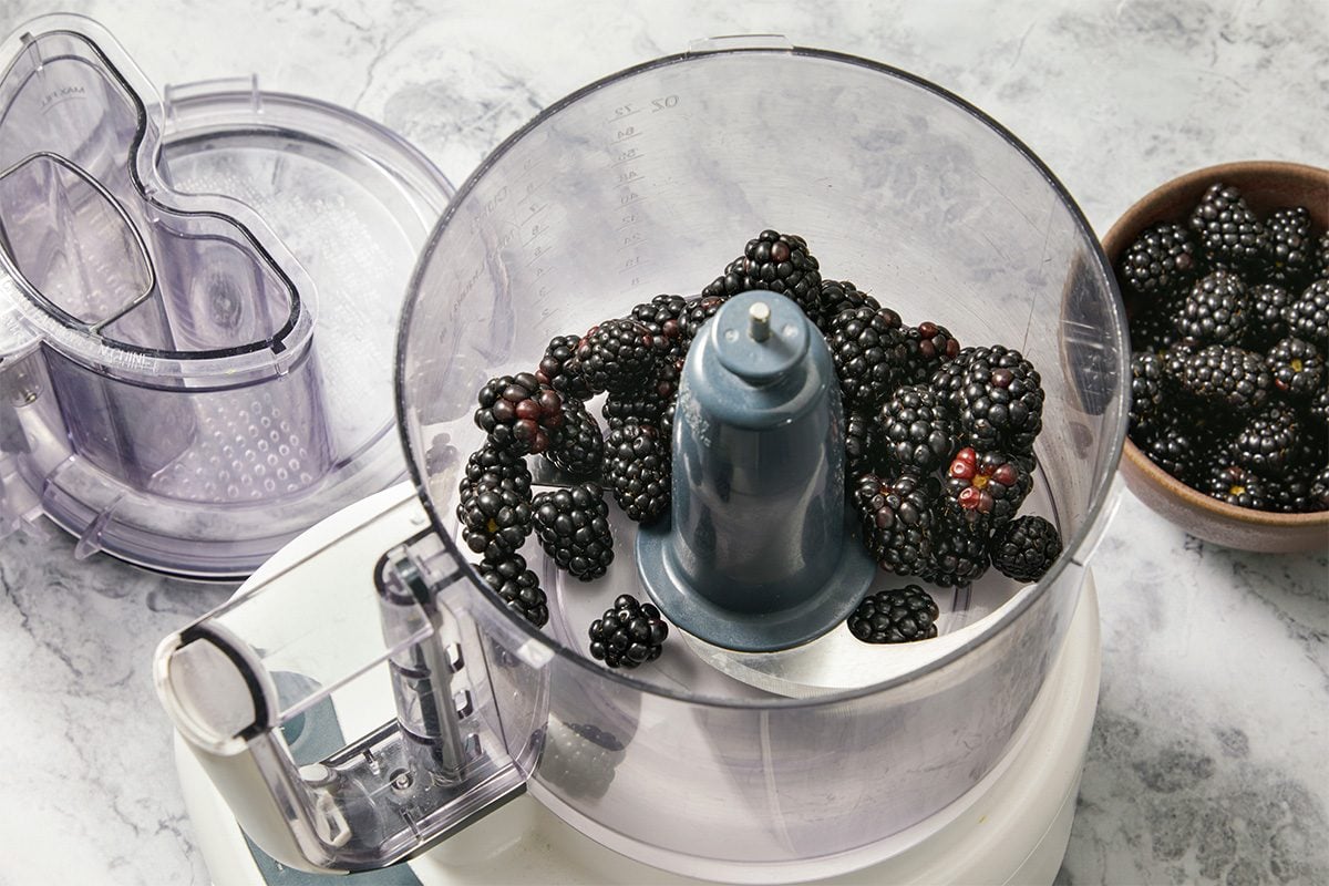 Eye Level horizontal image of a food processor with blackberries inside. A wooden bowl is shown with more blackberries beside the food processor, set on a marble countertop.