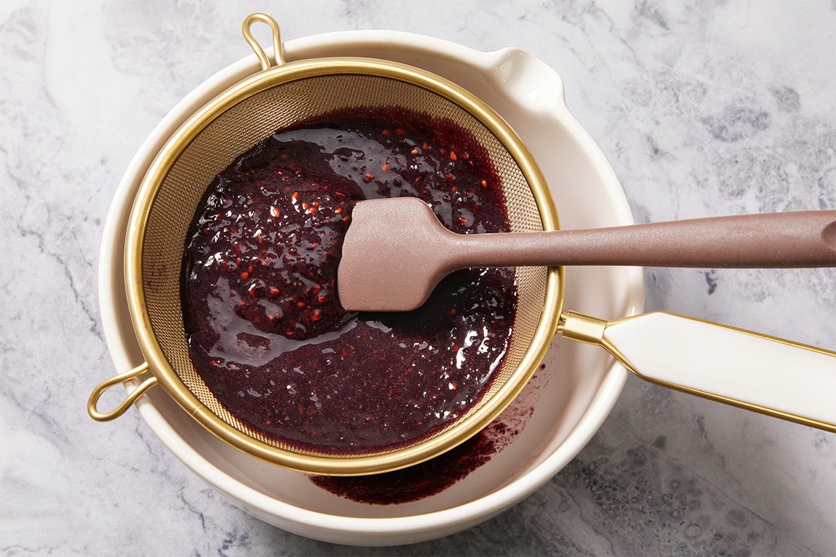 Overhead horizontal image of a strainer with dark red berry liquid being strained over a white bowl. A brown spatula rests inside the strainer.