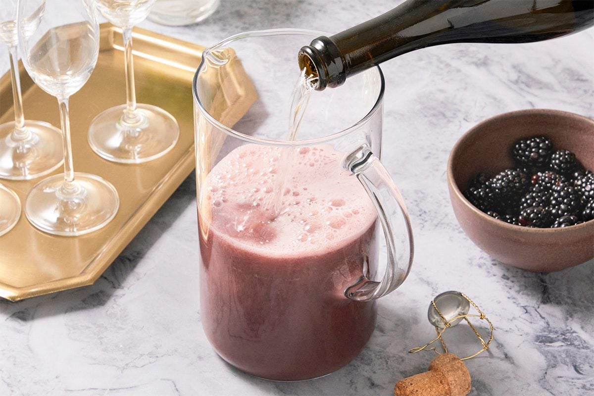 Eye Level horizontal image of sparkling wine being poured into a glass pitcher. A bowl of blackberries sits next to the pitcher, and Champagne flutes are on a gold tray in the background.