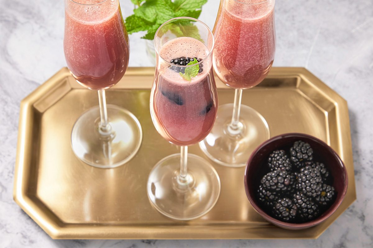 Overhead horizontal image of three champagne flutes filled with a red beverage, garnished with blackberries and mint sprigs, set on a gold tray next to a bowl of blackberries. The drink is a cool red color, and the blackberries are fresh and glossy.