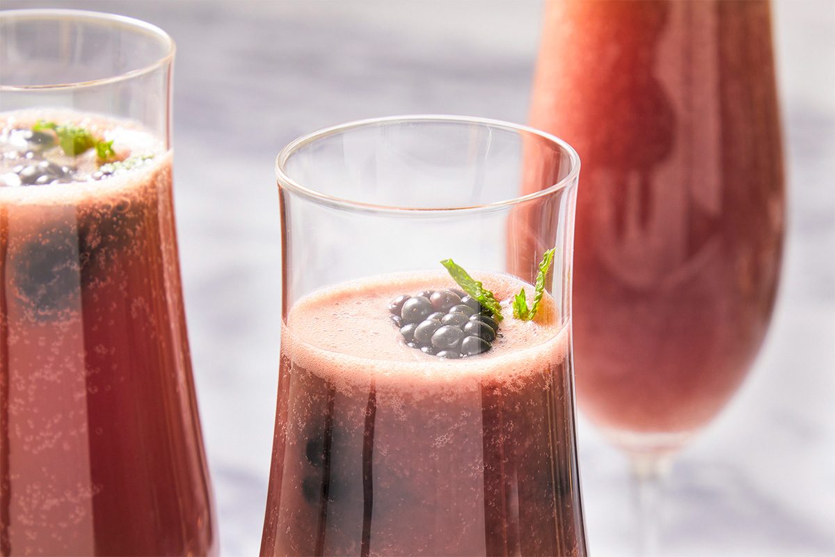 Eye Level horizontal image of three glasses filled with a blended red liquid, garnished with blackberries and mint sprigs. Bubbles are visible throughout the beverage and on the surface.