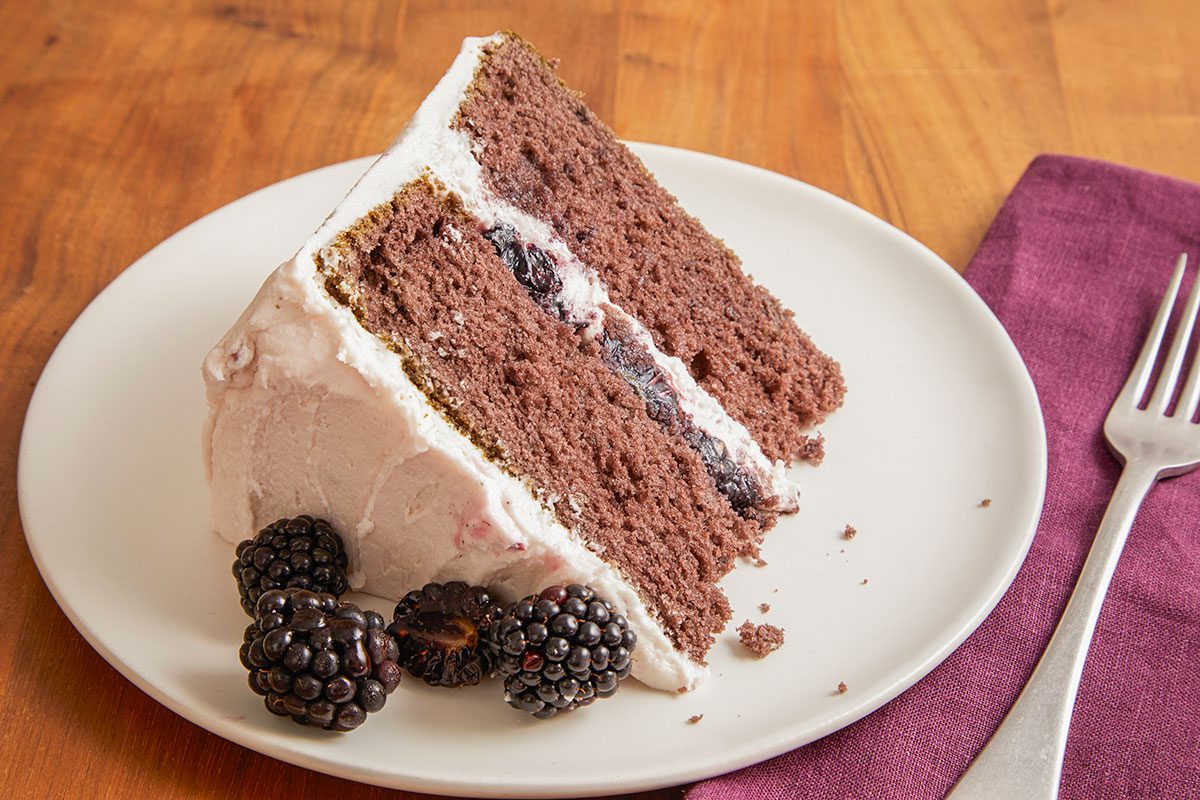 A horizontal image shows a slice of blackberry velvet cake on a plate with visible layers and frosting.