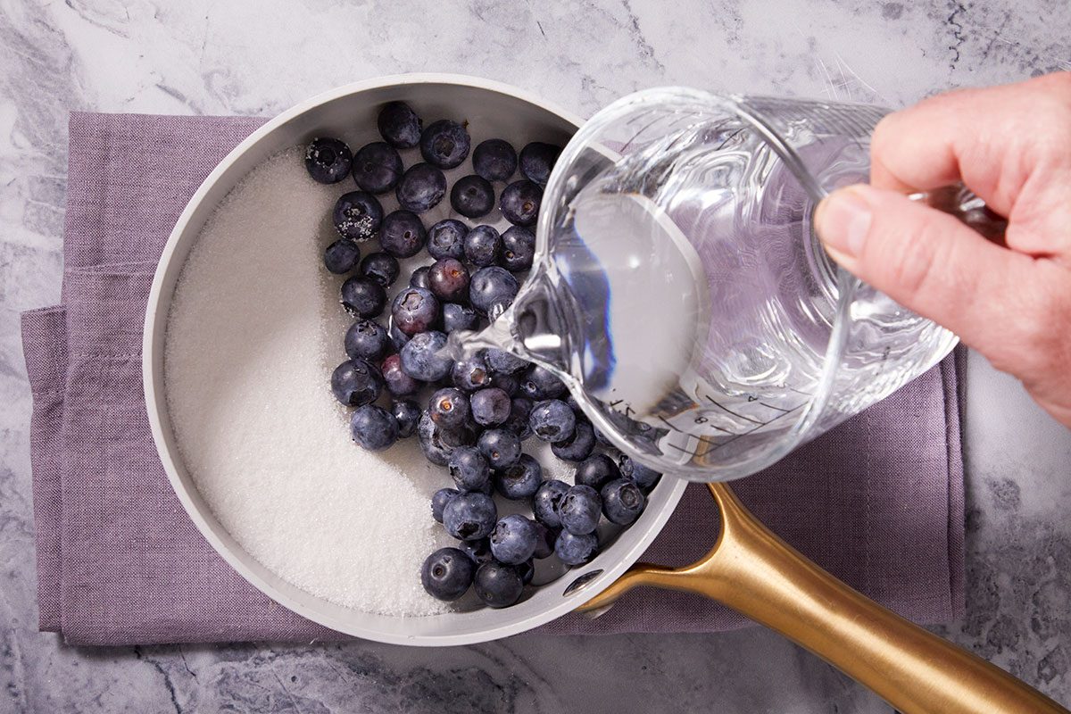 Overhead horizontal image of a white pan on a purple napkin, where blueberries and sugar have been placed in the pan. Water is being poured from a clear measuring cup into the pan.