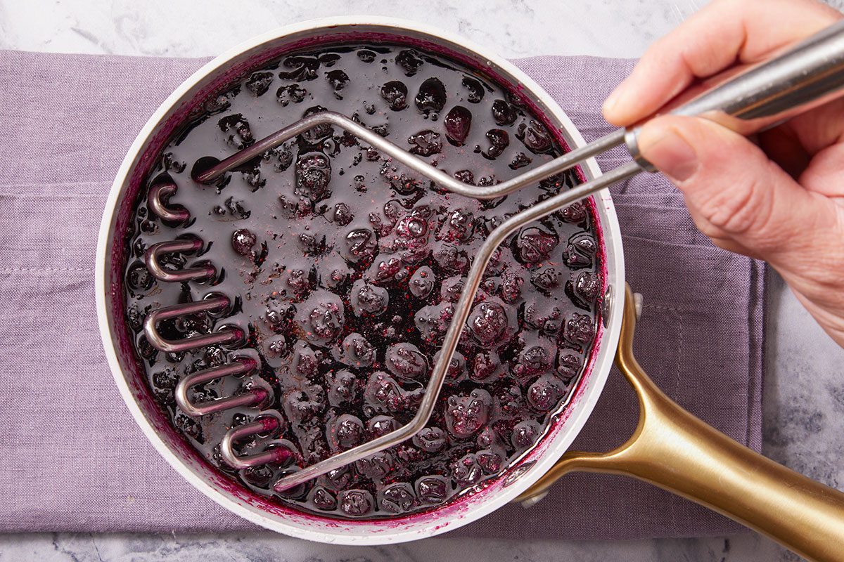 Overhead horizontal image of blueberries in a pot being mashed with a potato masher. The blueberries are dark purple and surrounded by a dark juice. A person's hand is visible holding the potato masher.
