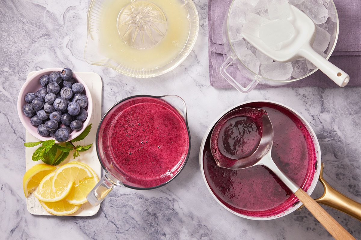 Overhead horizontal image of blueberry sauce being made. The image contains fresh blueberries, lemon slices, lemon juice, ice, and a pot with blueberry sauce and ladle.