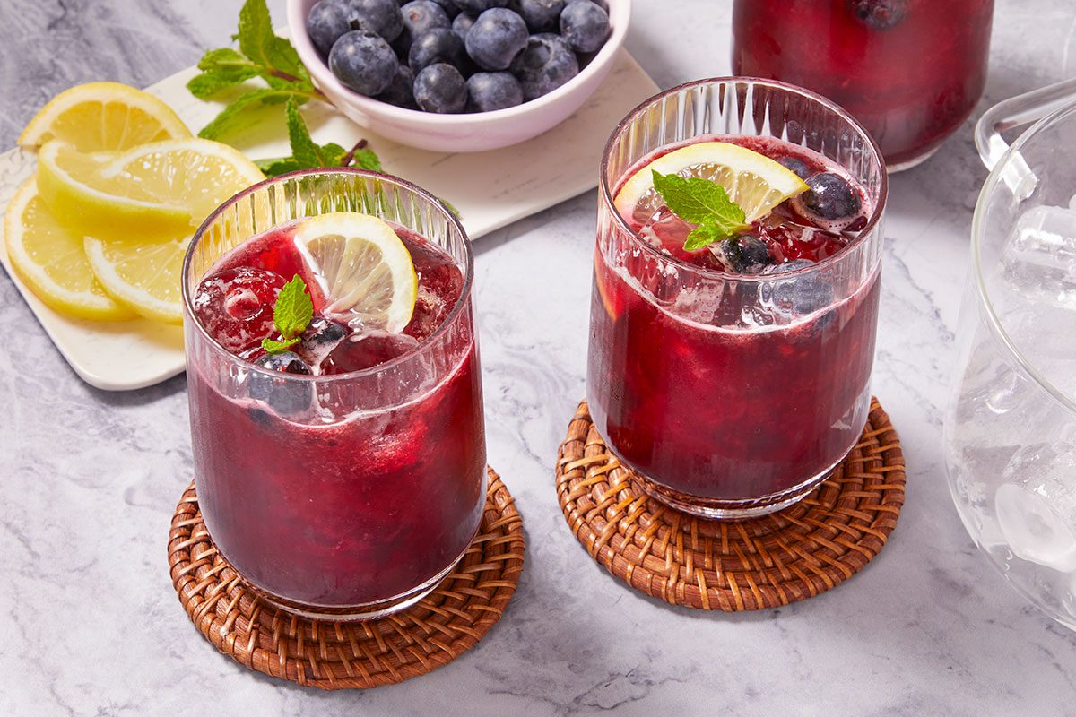 Eye level horizontal image of blueberry lemonade in two glasses on wicker coasters. The drink is garnished with lemon slices and mint. A bowl of blueberries, sliced lemons and ice are also in the frame.
