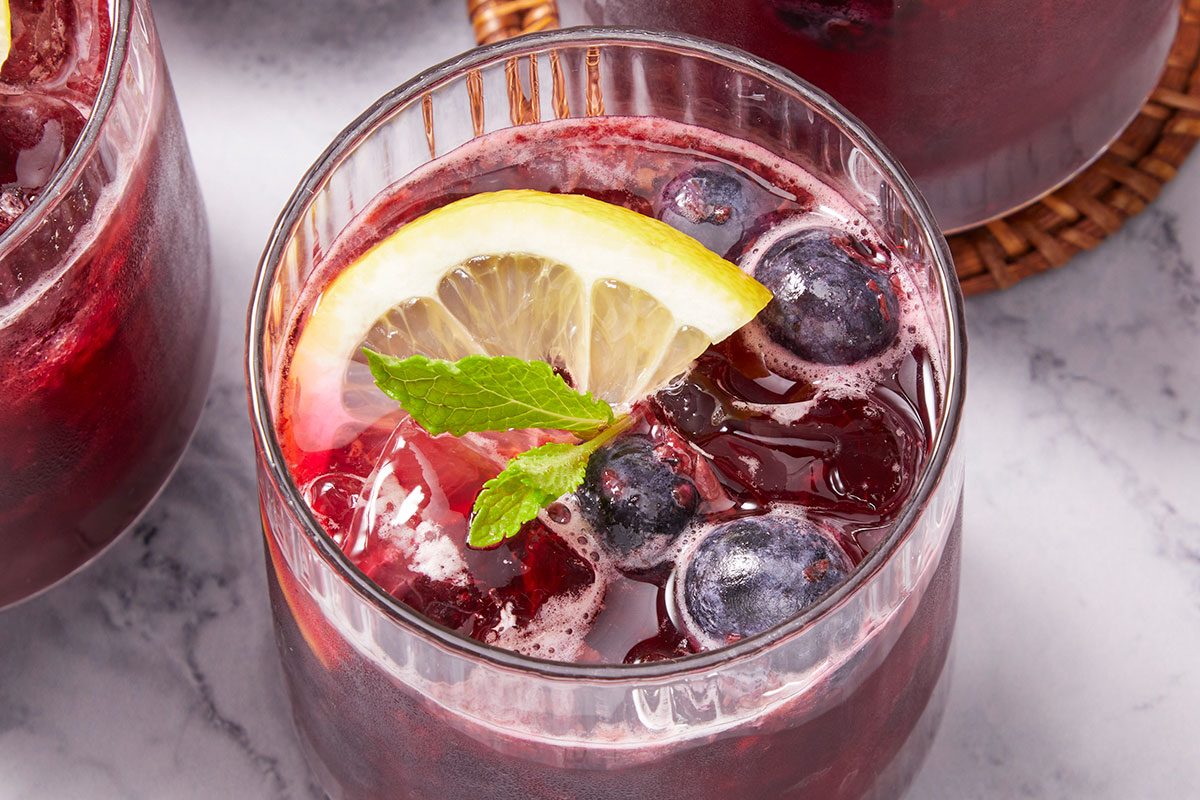 Overhead horizontal image of glasses filled with a deep red beverage, garnished with lemon slices, mint leaves, and blueberries. Ice cubes are visible in the drinks.