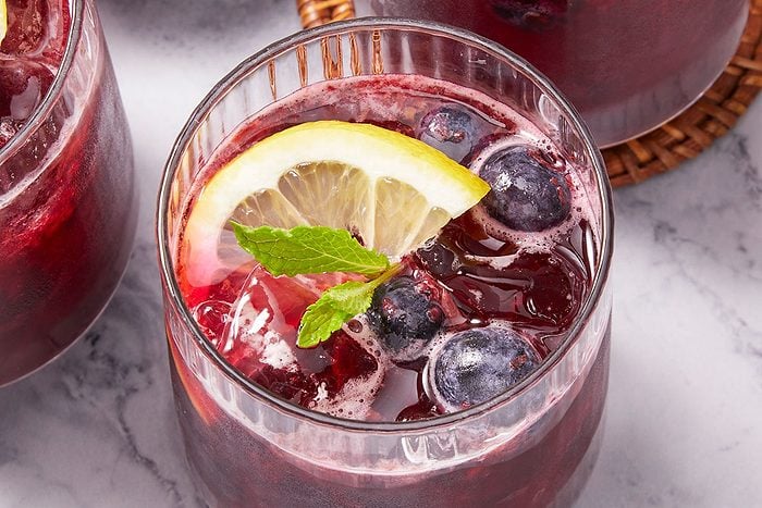 Overhead horizontal image of glasses filled with a deep red beverage, garnished with lemon slices, mint leaves, and blueberries. Ice cubes are visible in the drinks.