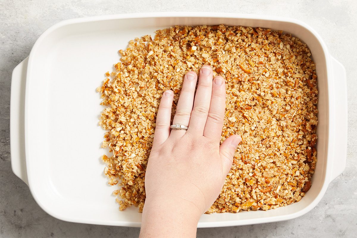 Overhead horizontal image of a Blueberry Pretzel Dessert step. A hand is pressing the crushed pretzels into a white baking dish. Visible ingredients include crushed pretzels, sugar, and butter.
