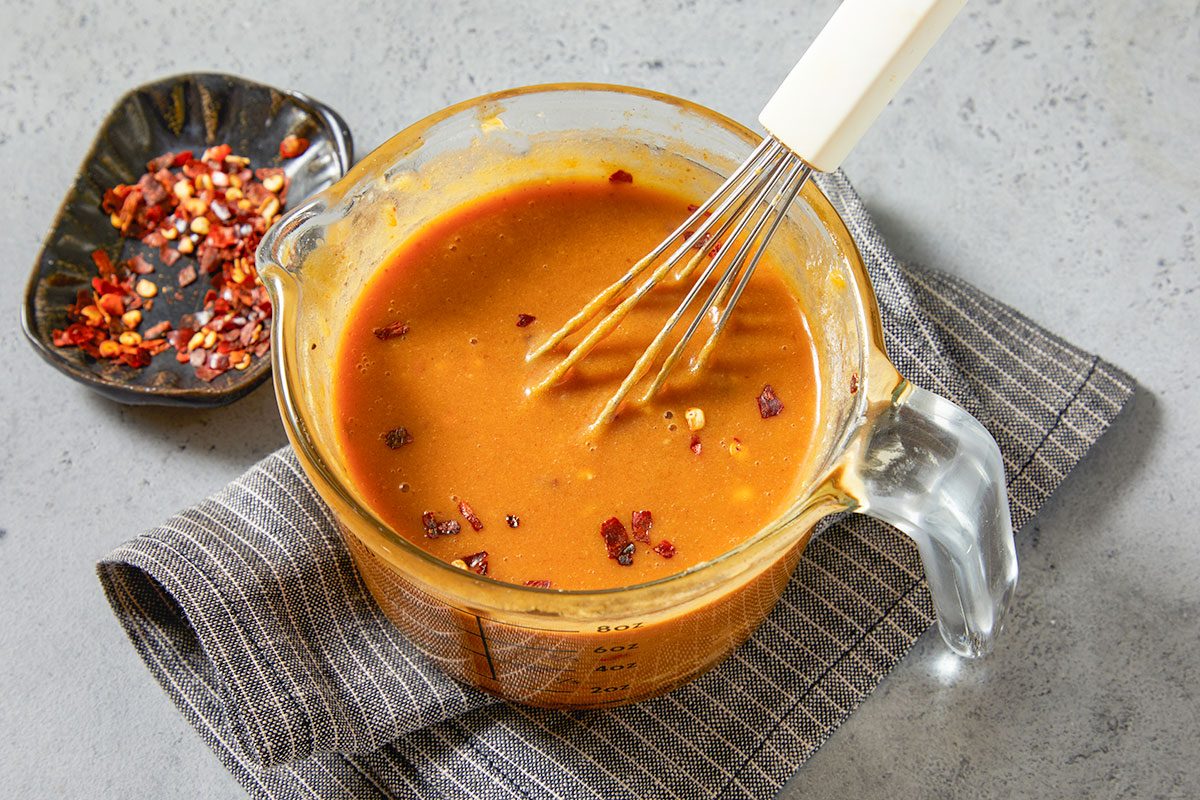 Overhead horizontal image of a clear measuring cup filled with a brown liquid mixture with flecks of red pepper flakes being whisked together. A small dish to the left contains more red pepper flakes.
