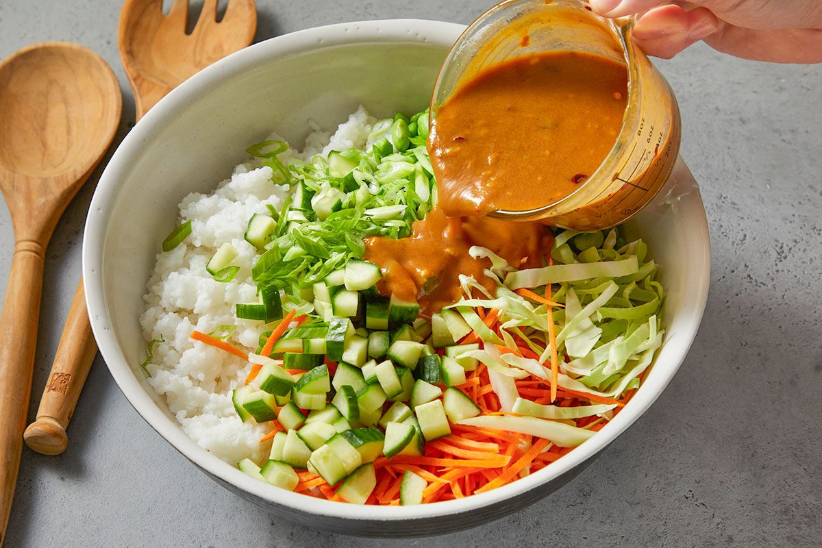 Eye Level horizontal image of a large bowl with rice, sliced cabbage, shredded carrots, diced cucumber, and chopped green onions. A hand is pouring a peanut sauce into the bowl from a clear glass measuring cup.