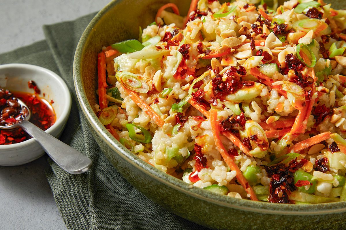 Eye Level horizontal image of rice salad in a bowl. The salad contains rice, carrots, sliced scallions, peanuts, cucumbers and is topped with a red chili crisp sauce.