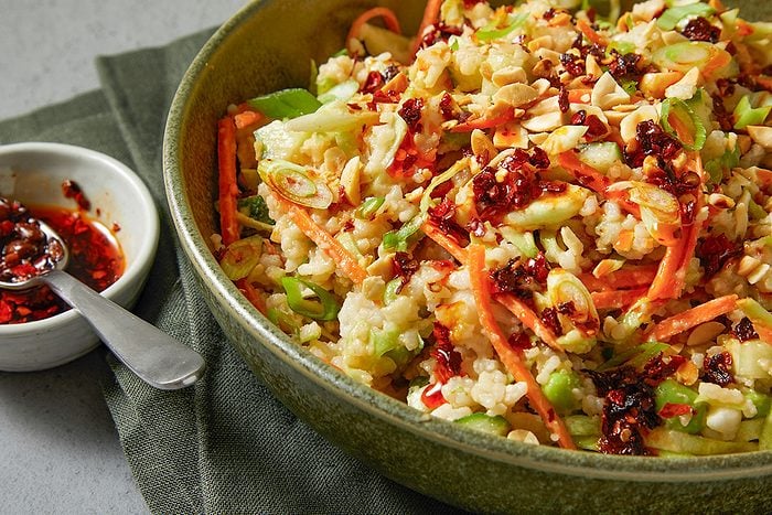 Eye Level horizontal image of rice salad in a bowl. The salad contains rice, carrots, sliced scallions, peanuts, cucumbers and is topped with a red chili crisp sauce.