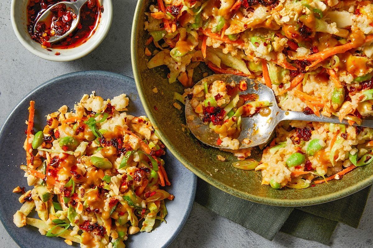 Overhead horizontal image of a plate and a bowl of savory food. The plate and bowl contain rice, carrots, peanuts, cabbage, edamame, and scallions. The bowl has a serving spoon in it and a small bowl of chili oil is next to it.