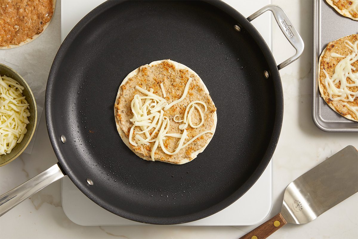 Overhead horizontal image of the recipe Elise Jesse's Smash Chicken Caesar Tacos. A flour tortilla is in a frying pan, topped with a chicken mixture and some shredded mozzarella cheese.
