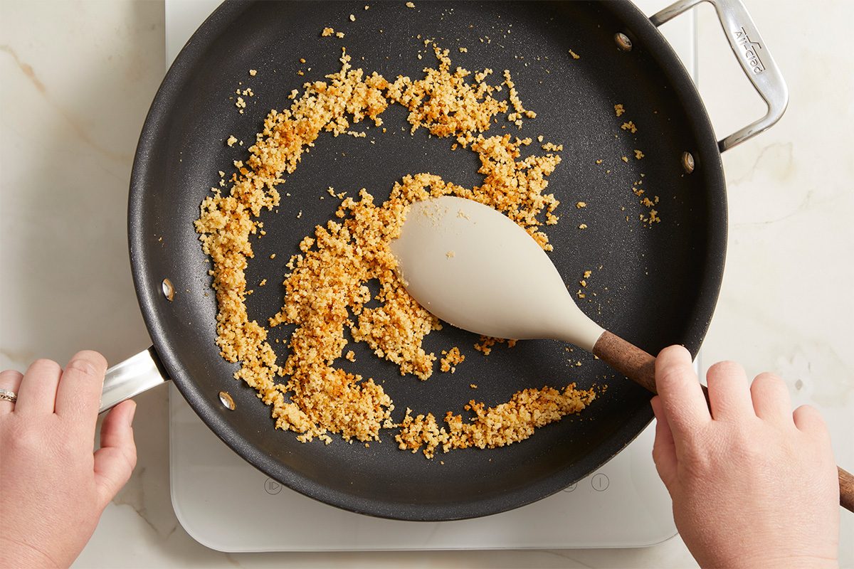 Overhead horizontal image of toasting breadcrumbs in a skillet. The breadcrumbs are golden brown, and a spatula is being used to stir them around in the pan.