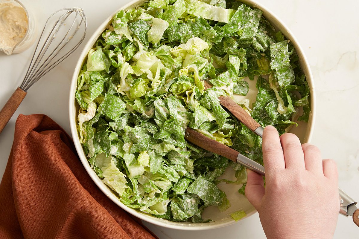 Overhead horizontal image of the romaine lettuce, tossed with the Caesar dressing, and Parmesan cheese in a bowl, using salad tongs. Also visible are a brown kitchen towel, a whisk, and the prepared Caesar dressing in a jar.