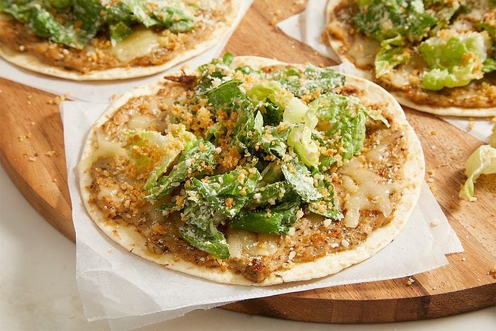 Eye Level horizontal image of several Smash Chicken Caesar Tacos on a wooden board. The tacos are topped with ground chicken, melted mozzarella cheese, romaine lettuce, Caesar dressing, grated Parmesan cheese, and dry bread crumbs.