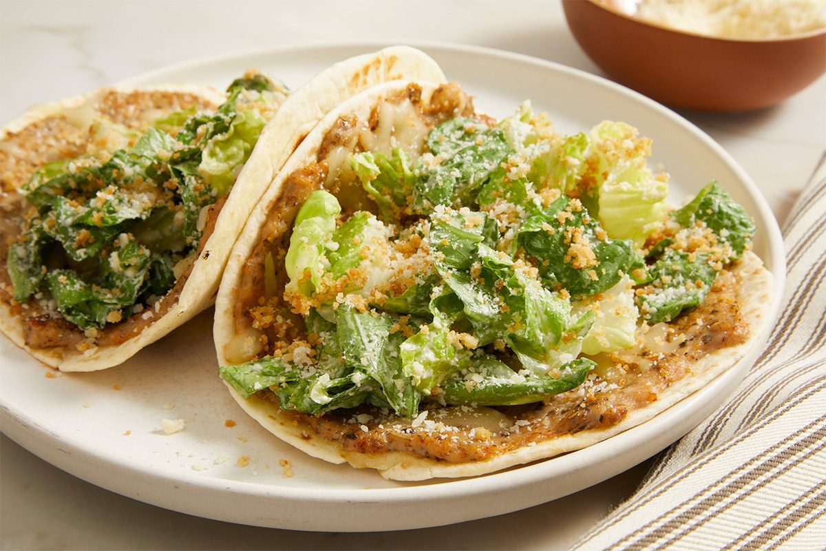 Eye level horizontal image of Elise Jesse's Smash Chicken Caesar Tacos. Two tacos rest on a white plate, filled with seasoned ground chicken, shredded mozzarella cheese, romaine lettuce, grated Parmesan cheese, and dry bread crumbs.