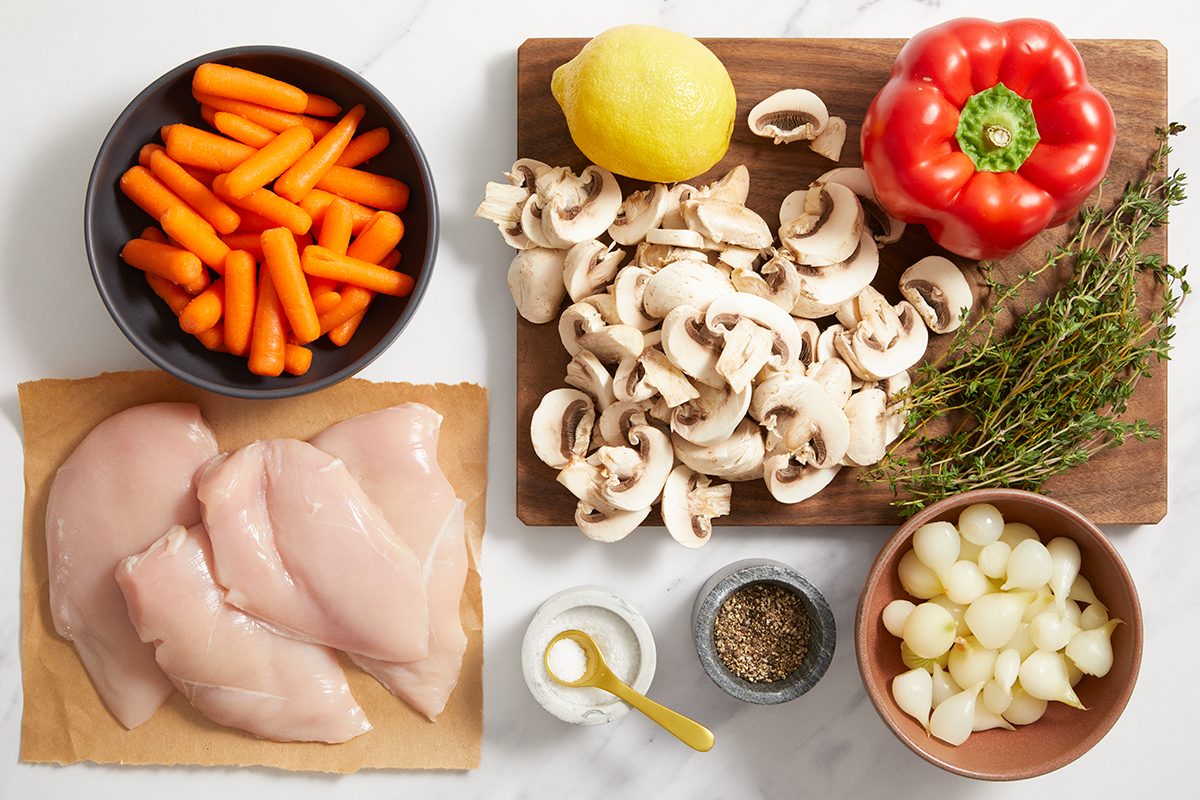 Raw chicken breasts, baby carrots, pearl onions, sliced mushrooms, a red bell pepper, a lemon, fresh thyme sprigs, salt, pepper, and a small bowl of flour are arranged on a white surface and wooden board.