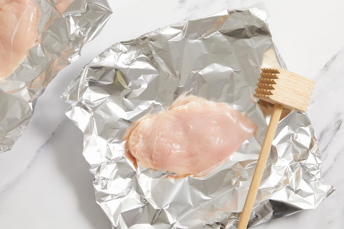 Raw chicken breast placed on a sheet of aluminum foil, with a wooden meat tenderizer lying next to it on a white marble surface.