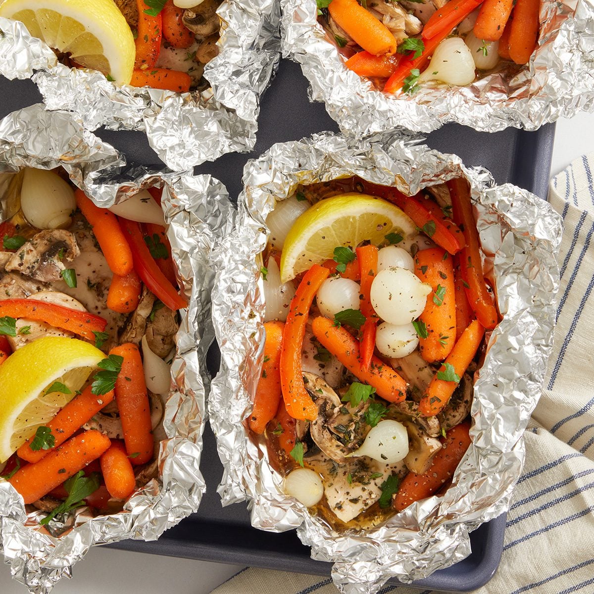 Four foil packets filled with seasoned chicken, baby carrots, pearl onions, red bell peppers, and lemon slices are arranged on a baking tray, ready for cooking. A striped cloth is partially visible beside the tray.