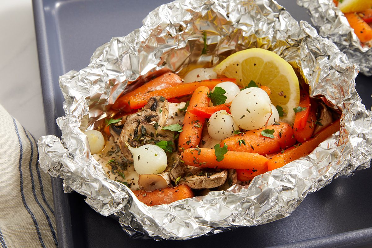 A foil packet filled with cooked baby carrots, pearl onions, mushrooms, herbs, parsley, and a slice of lemon, placed on a dark tray.