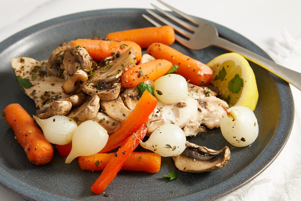 A plate of cooked chicken breast topped with mushrooms, pearl onions, baby carrots, and red bell pepper strips, garnished with herbs, with a lemon wedge and fork on the side.