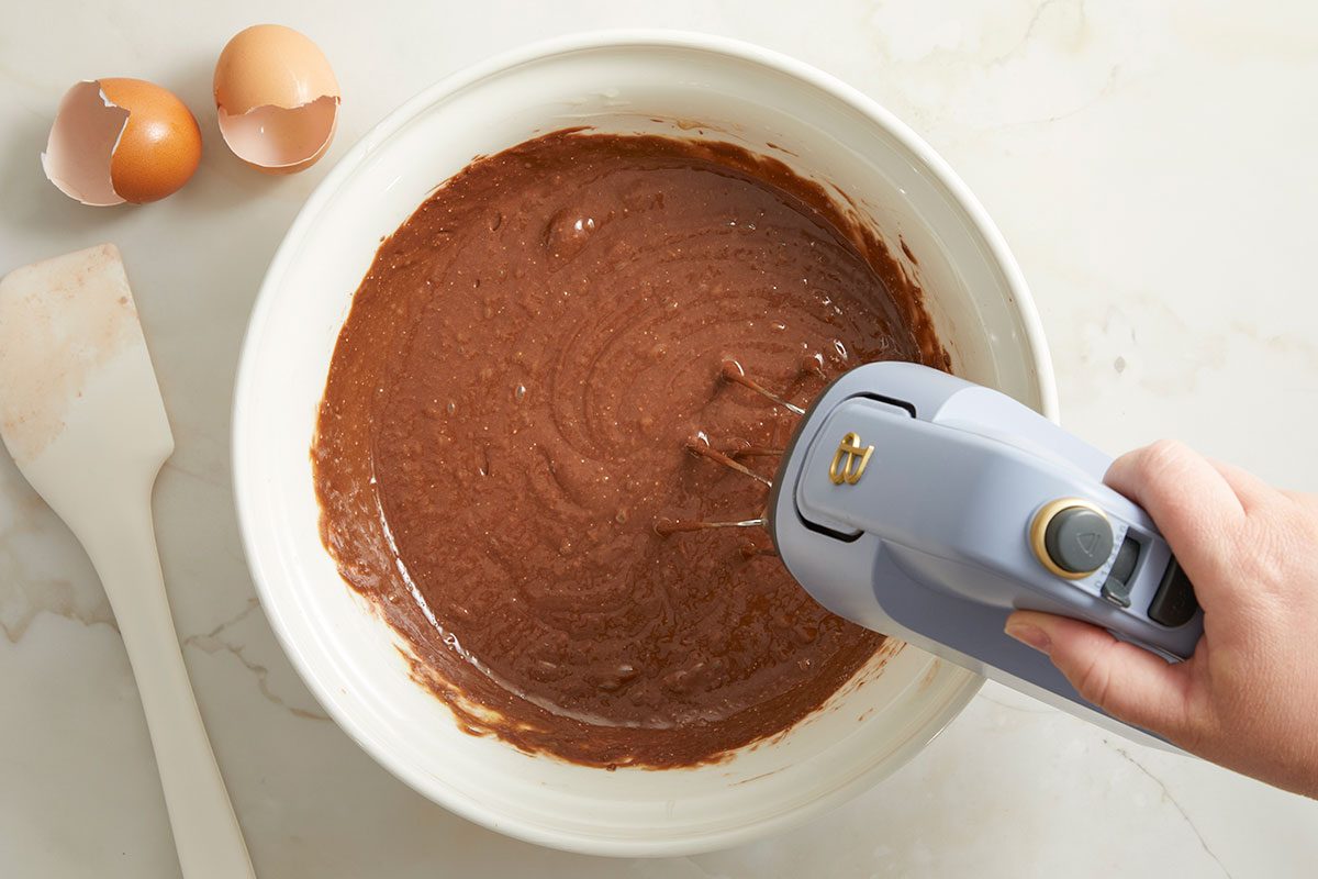 Overhead horizontal image of a chocolate batter being mixed with a handheld electric mixer. The batter is in a white bowl and there are two empty eggshells and a spatula next to the bowl.