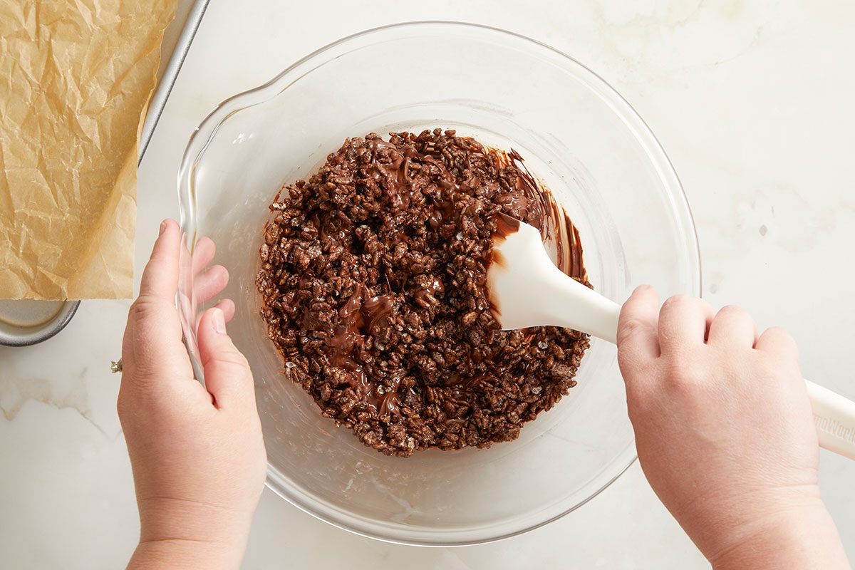 Overhead horizontal image of hands mixing chocolate into crispy rice cereal in a large glass bowl with a white spatula. In the background is a baking sheet lined with parchment paper.
