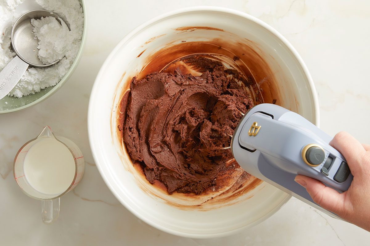 Overhead horizontal image of a Step by Step shot mixing chocolate batter in a white mixing bowl with a handheld electric mixer. A measuring cup filled with powdered sugar and a glass measuring pitcher filled with milk sit to the side.
