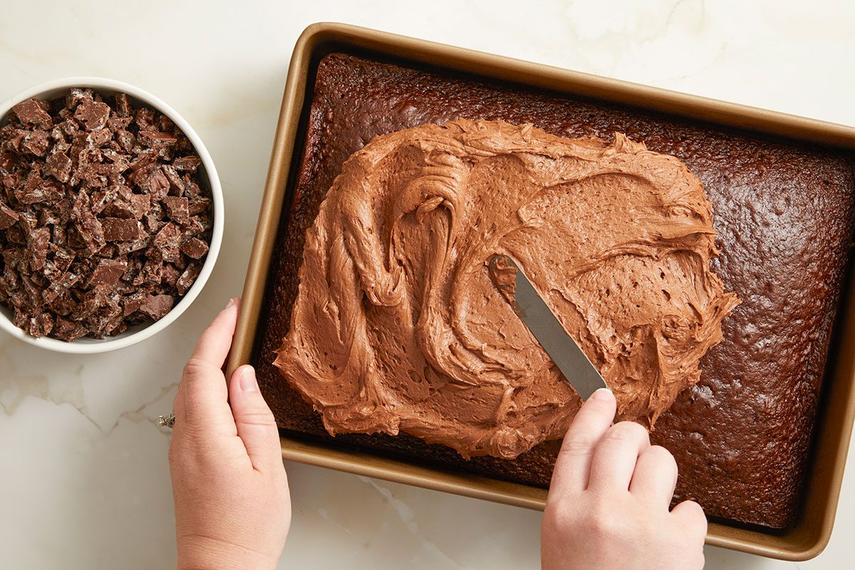 Overhead horizontal image of applying chocolate frosting to a chocolate cake in a metal baking pan with an offset spatula. A bowl of chocolate chunks is visible next to the cake.