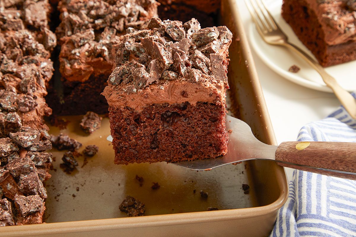 Eye Level horizontal image of a slice of chocolate cake being lifted from the baking pan with a spatula. The cake has a thick layer of chocolate frosting and is topped with chopped chocolate candy pieces.