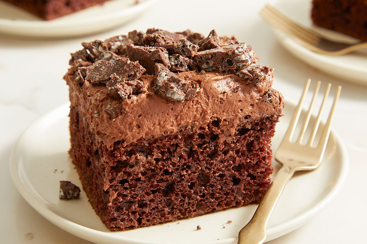 Eye Level horizontal image of a slice of chocolate cake on a white plate with a gold fork. The cake is topped with chocolate frosting and chunks of chocolate.