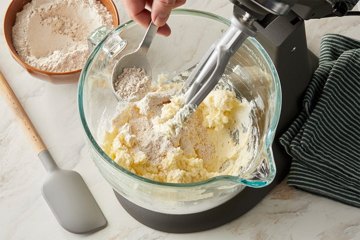 Eye Level horizontal image of a mixing bowl with dough being prepared in a stand mixer. A hand is pouring a scoop of flour into the bowl. A spatula is also visible.