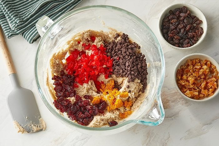 Overhead horizontal image of cookie dough in a glass bowl with red glace cherries, dark chocolate chips, dried cranberries and candied orange peel. Raisins and diced candied ginger are in separate bowls next to the bowl of cookie dough.