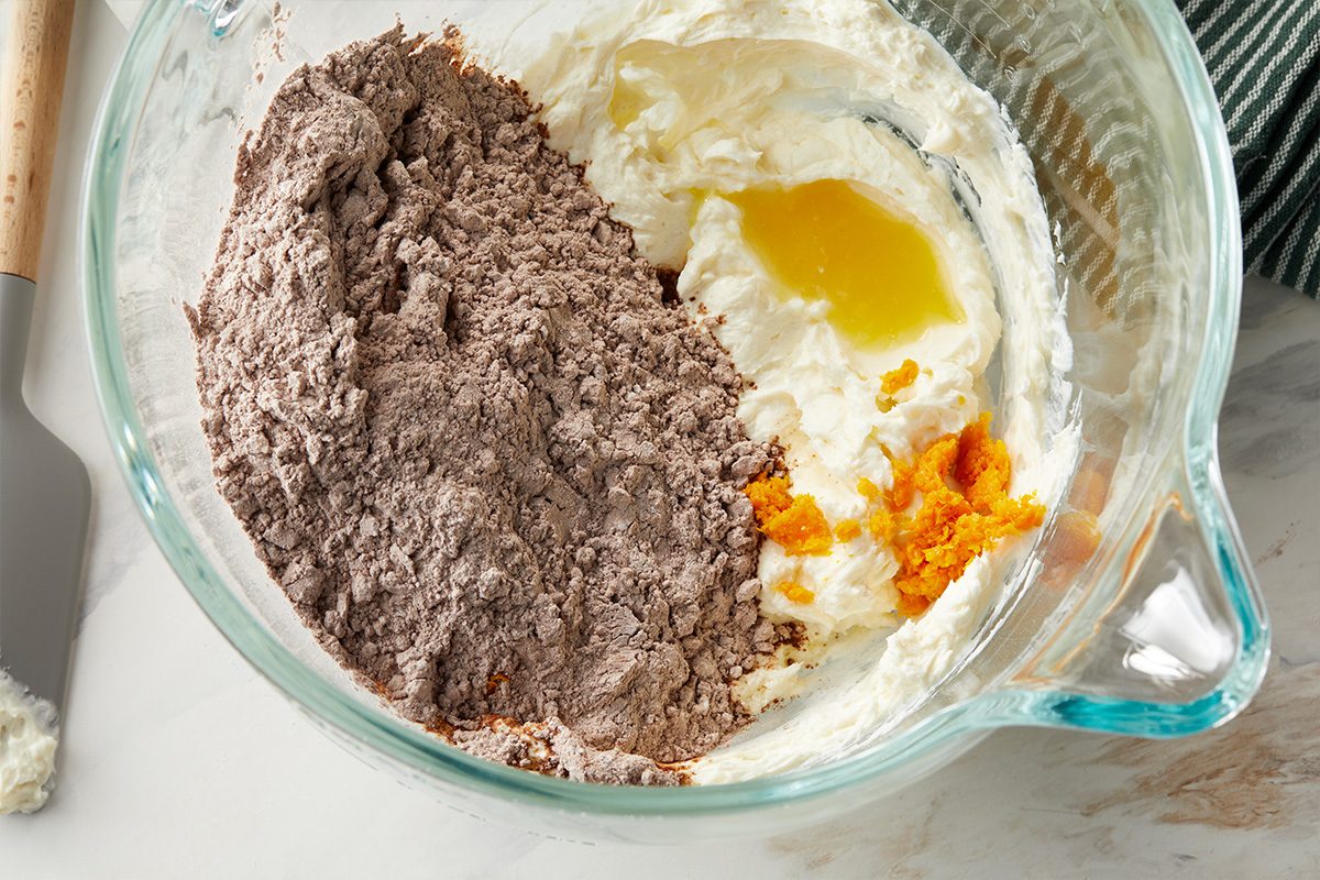 Overhead horizontal image of the ingredients in a clear mixing bowl. The bowl contains a pile of chocolate powder, a mass of white cream cheese, a pool of melted butter, and a pile of orange zest. A gray spatula rests on the side.
