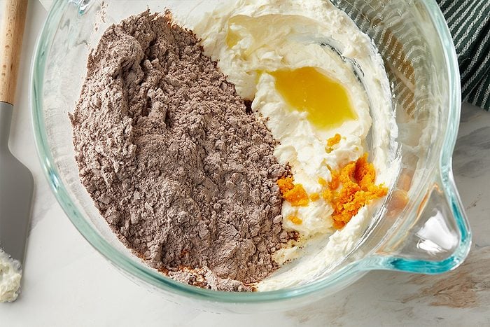 Overhead horizontal image of the ingredients in a clear mixing bowl. The bowl contains a pile of chocolate powder, a mass of white cream cheese, a pool of melted butter, and a pile of orange zest. A gray spatula rests on the side.