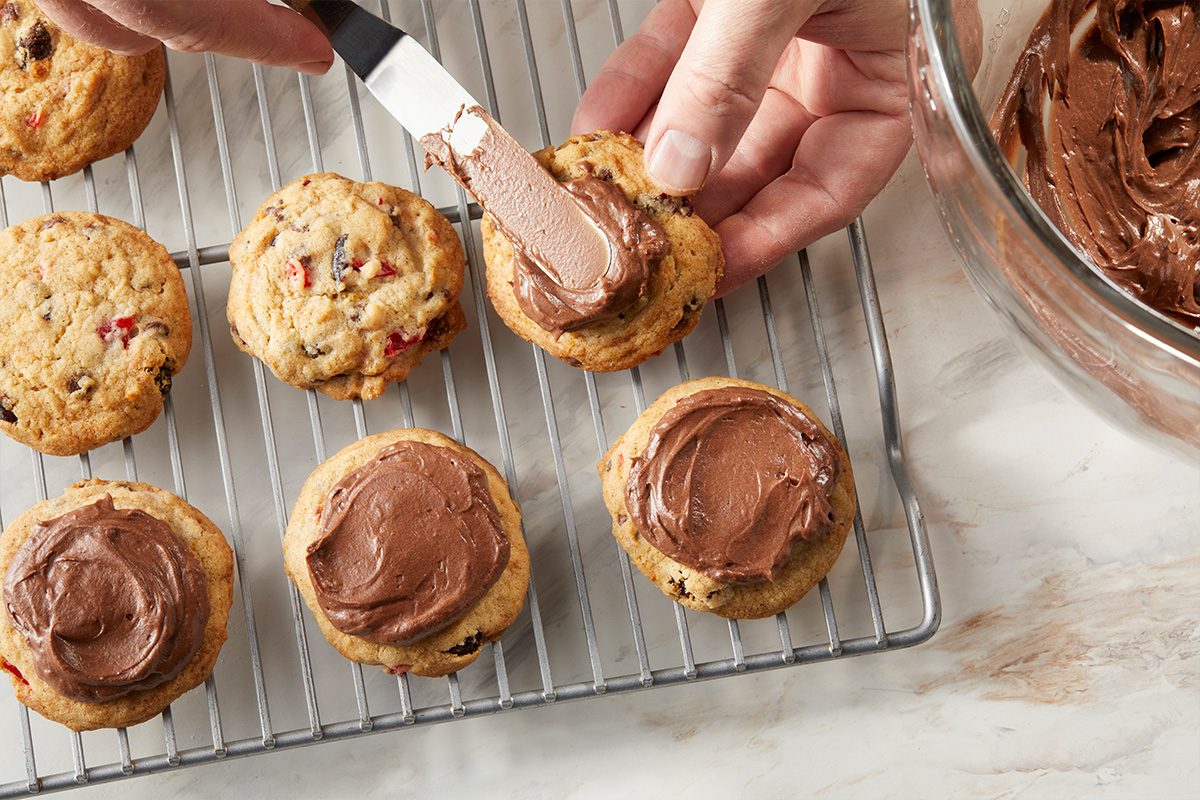 Overhead horizontal image of a hand using a spatula to spread chocolate frosting on a cookie. Several other cookies are visible on a wire cooling rack and a glass bowl of chocolate frosting is on the side.